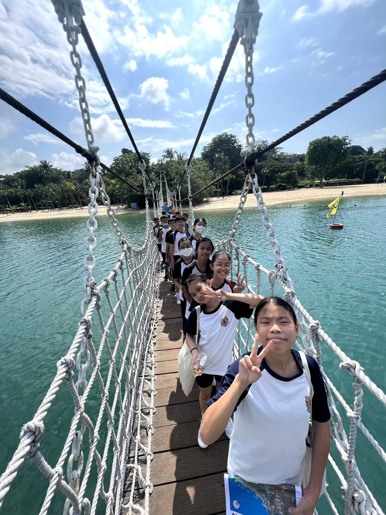A line of students in school uniforms walks across a rope suspension bridge over turquoise water near a sandy beach.